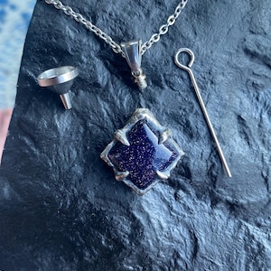 May include: A silver necklace with a blue stone pendant, a small funnel, and a tool. The pendant is a square shape with a dark blue, glittery centre. The items are displayed on a dark, textured surface.