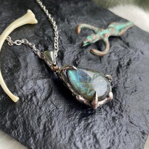 May include: A silver pendant necklace with a teardrop-shaped labradorite stone. The stone has blue and green iridescent flashes. A curved bone and a small bronze figure are also present on a textured black surface.