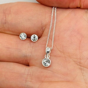 May include: A silver necklace and earrings set with clear crystal stones. The necklace has a simple chain and a pendant with a single round crystal.