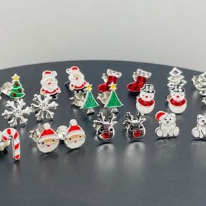 Puede incluir: Una colección de 18 pendientes de botón de temática navideña plateados. Los pendientes presentan varios diseños, incluyendo Papá Noel, renos, muñecos de nieve, bastones de caramelo, copos de nieve y árboles de Navidad. Los pendientes son todos plateados con detalles de esmalte de colores.