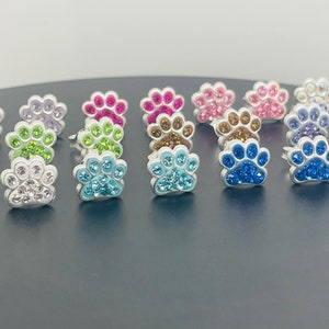 May include: A collection of 18 pairs of silver paw print earrings with various colored crystal accents. The earrings are arranged in a row on a dark gray surface.