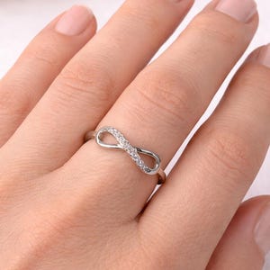 Puede incluir: Un anillo plateado con forma de símbolo de infinito, adornado con pequeñas piedras preciosas transparentes. El anillo se muestra en una mano, mostrando su delicado diseño y brillo. El anillo es una joya.