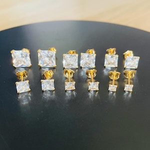 May include: An assortment of gold-tone square stud earrings, each featuring a clear, square-cut gemstone. The earrings are presented in rows, with sizes ranging from large to small. The studs are displayed on a dark surface.