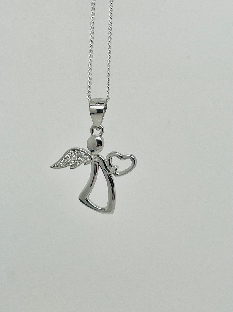 以下が含まれることがあります： ハートのチャームが付いたシルバーのエンジェルペンダントネックレス。エンジェルは小さなハローと、きらめく石のディテールが施された翼を持っています。