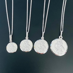 Puede incluir: Cuatro collares con colgantes de San Cristóbal de plata, con colgantes redondos de diferentes tamaños. Cada colgante presenta una imagen grabada de San Cristóbal y cuelga de una fina cadena de plata. Sobre un fondo oscuro.