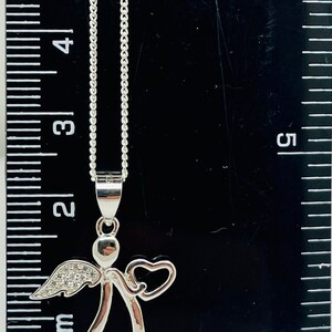 以下が含まれることがあります： ハートとクリアクリスタルアクセントが付いたシルバーエンジェルペンダントネックレス。ネックレスの長さは45.7cmで、定規の上に置かれています。