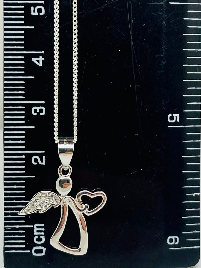 以下が含まれることがあります： ハートとクリアクリスタルアクセントが付いたシルバーエンジェルペンダントネックレス。ネックレスの長さは45.7cmで、定規の上に置かれています。