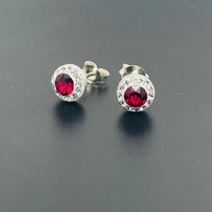 Peut inclure: Une paire de boucles d'oreilles en argent avec un centre en cristal rouge entouré d'un halo de petits cristaux blancs.