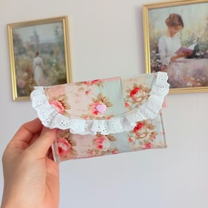 May include: A floral print wallet with a pink heart-shaped button closure. The wallet features a white lace trim and a patchwork design with pink and blue floral patterns. The wallet is held in a hand.