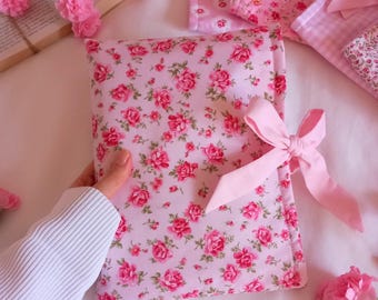 May include: A pink floral print pillow with a pink satin ribbon tied around it. The pillow is on a white surface with pink flowers.