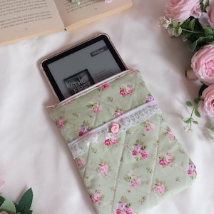 May include: A quilted, sage green e-reader sleeve with a floral pattern of pink and purple roses. The sleeve has a white lace trim and a small pink rose accent. An e-reader is partially visible inside the sleeve.