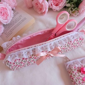 May include: A floral print pencil case with a white lace trim and a pink bow. Inside, pink scissors and a pen are visible. The case is open, showing a pink interior. Pink roses and a book are in the background.