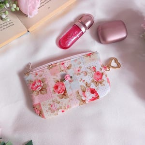 以下が含まれることがあります： 金色のハート型のチャームが付いた花柄の小銭入れ、ピンクのリップグロス、小さなピンクのケース。小銭入れはピンクのバラと小さなバラの装飾が施されたパッチワークデザインです。アイテムは白い表面に配置されています。