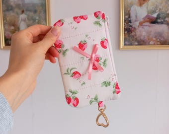 May include: A white quilted wallet with a strawberry pattern, a pink bow, and a gold heart-shaped charm. The wallet has a zipper closure and is held up by a hand.
