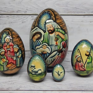 Peut inclure: Un ensemble de cinq œufs en bois peints à la main représentant des scènes de la nativité. Le plus grand œuf illustre la sainte famille, tandis que les autres montrent les rois mages, des bergers, un agneau et une colombe sous un ciel étoilé.