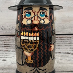 May include: A hand-painted wooden doll depicting a man with a black hat, glasses, and a long beard. The doll holds a menorah with lit candles. The doll's clothing is painted in shades of gray and brown.