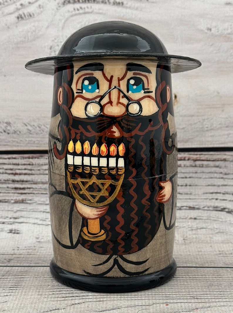 May include: A hand-painted wooden doll depicting a man with a black hat, glasses, and a long beard. The doll holds a menorah with lit candles. The doll's clothing is painted in shades of gray and brown.