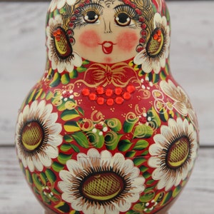 May include: A hand-painted Russian nesting doll, or Matryoshka doll, with a red base color. The doll features a painted face, floral designs, and gold accents. The flowers are white with yellow centers.