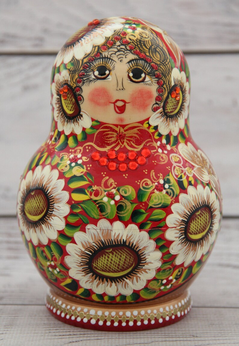 May include: A hand-painted Russian nesting doll, or Matryoshka doll, with a red base color. The doll features a painted face, floral designs, and gold accents. The flowers are white with yellow centers.