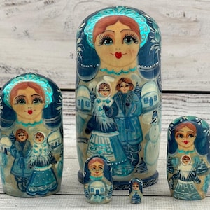 Può includere: Un set di cinque bambole matrioska con tema invernale nei toni del blu, bianco e argento. Ogni bambola presenta un volto dipinto con labbra rosse e una scena invernale con figure in cappotto, case innevate e un pupazzo di neve.