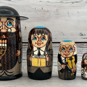 May include: A set of five hand-painted wooden nesting dolls. The largest doll depicts a man with a black hat and beard holding a menorah. Other dolls show figures with books, a violin, and bread. The dolls are painted in shades of brown, black, and beige.