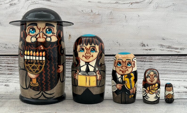 May include: A set of five hand-painted wooden nesting dolls. The largest doll depicts a man with a black hat and beard holding a menorah. Other dolls show figures with books, a violin, and bread. The dolls are painted in shades of brown, black, and beige.