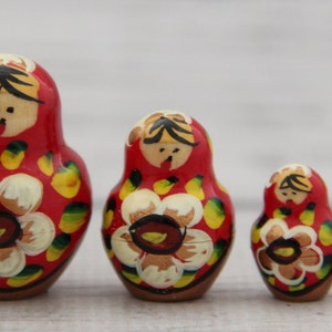 May include: A set of four red Matryoshka nesting dolls. Each doll features a painted floral design with yellow and white petals. The dolls have a wooden appearance and are arranged from largest to smallest.