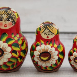 May include: Three traditional Russian nesting dolls, or Matryoshka dolls, stand in a row. Each doll is painted red with a floral design featuring white and yellow flowers and green leaves. The dolls have painted faces.