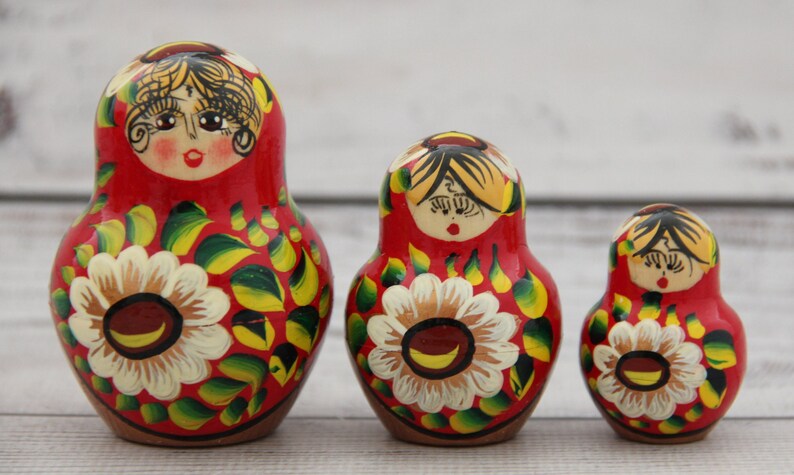 May include: Three traditional Russian nesting dolls, or Matryoshka dolls, stand in a row. Each doll is painted red with a floral design featuring white and yellow flowers and green leaves. The dolls have painted faces.