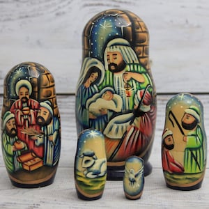 May include: A set of six hand-painted wooden nesting dolls depicting a Nativity scene. The dolls feature detailed illustrations of Mary, Joseph, baby Jesus, the Three Wise Men, and a shepherd. The color palette includes blues, greens, and browns.