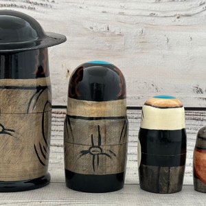 May include: A set of five wooden nesting dolls, each with a unique design. The largest doll has a black hat and a light brown body with black accents. The other dolls have black, brown, and teal details. The dolls are arranged in a row.
