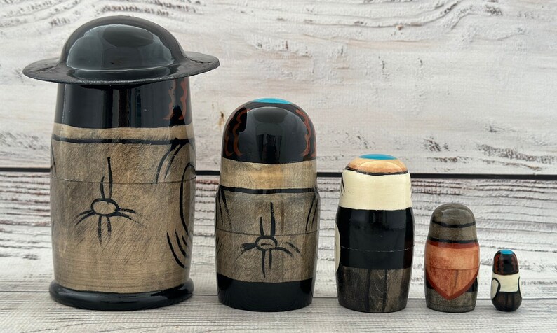 May include: A set of five wooden nesting dolls, each with a unique design. The largest doll has a black hat and a light brown body with black accents. The other dolls have black, brown, and teal details. The dolls are arranged in a row.