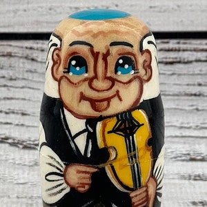 May include: A hand-painted wooden nesting doll depicting an elderly man with a blue cap, white hair, and a black suit. He is holding a yellow violin. The doll has a smiling face with blue eyes.