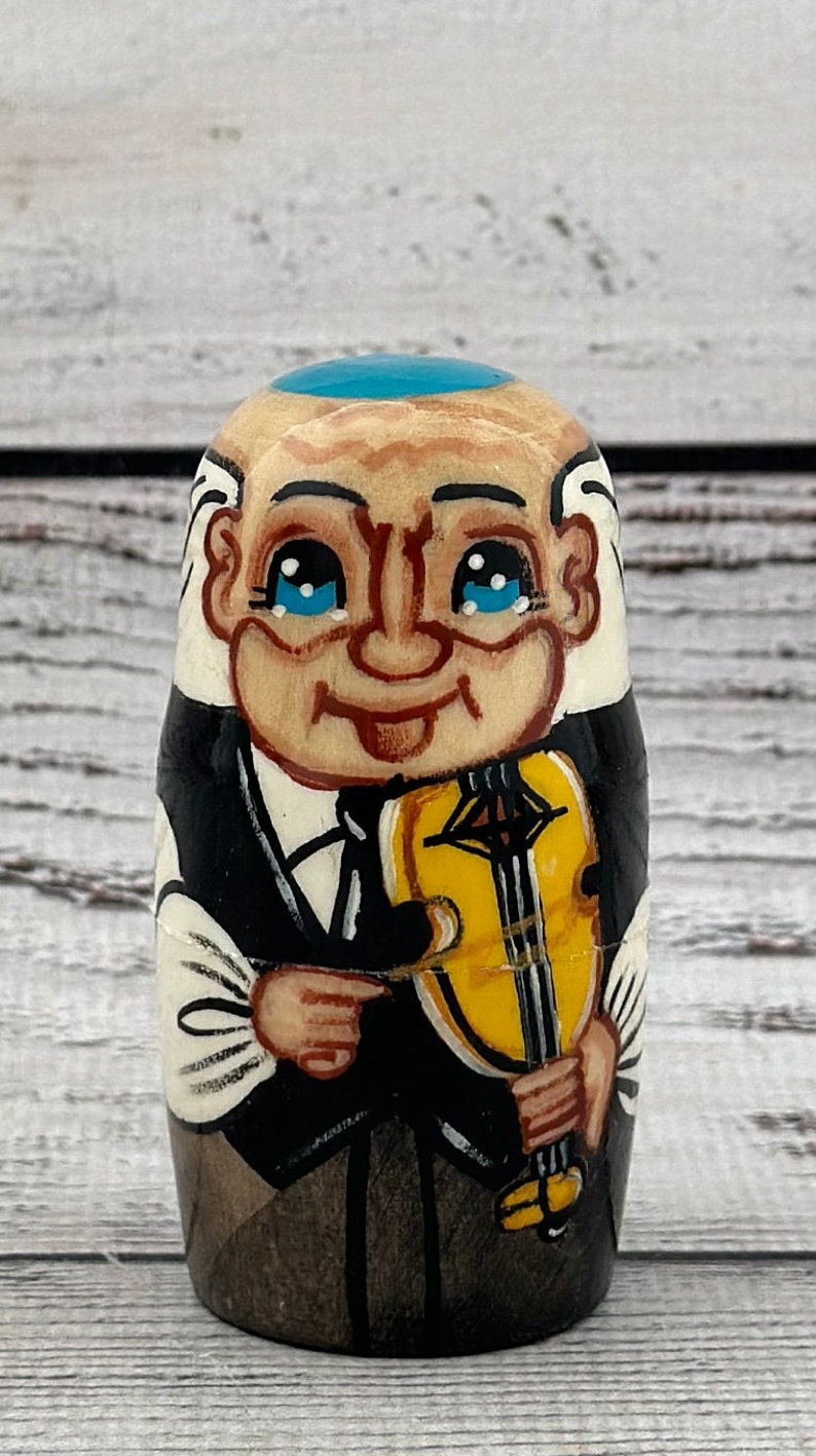 May include: A hand-painted wooden nesting doll depicting an elderly man with a blue cap, white hair, and a black suit. He is holding a yellow violin. The doll has a smiling face with blue eyes.