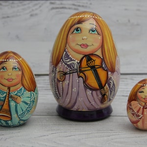 Peut inclure: Trois poupées russes en bois peintes à la main en forme d'œuf. Les poupées ont des cheveux blonds, des joues roses et des vêtements détaillés. La plus grande poupée tient un violon, la poupée du milieu tient une trompette et la plus petite poupée a un col décoratif.