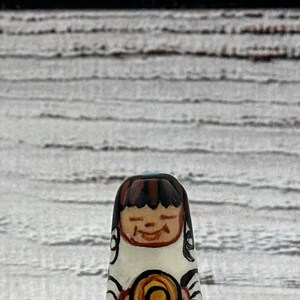 May include: A hand-painted wooden doll with a smiling face, brown hair, and a black base. The doll is holding a yellow and orange circular object. The doll is approximately 3 inches tall.