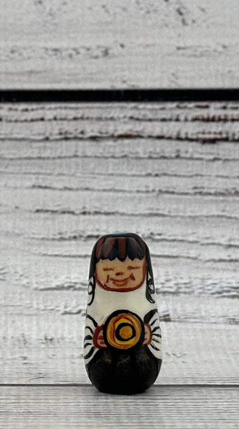 May include: A hand-painted wooden doll with a smiling face, brown hair, and a black base. The doll is holding a yellow and orange circular object. The doll is approximately 3 inches tall.