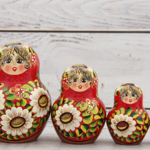May include: A set of traditional Russian nesting dolls, or Matryoshka dolls, in varying sizes. Each doll features a red base with floral and facial details. The largest doll is approximately 6 inches tall.