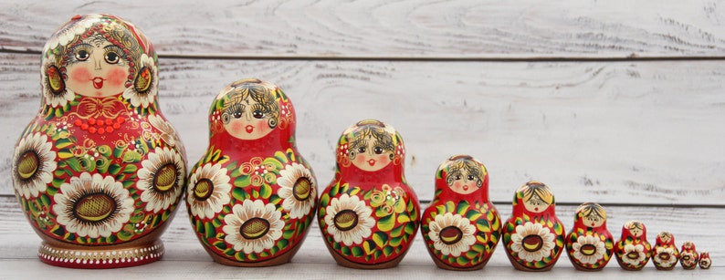 May include: A set of traditional Russian nesting dolls, or Matryoshka dolls, in varying sizes. Each doll features a red base with floral and facial details. The largest doll is approximately 6 inches tall.