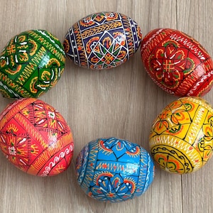 May include: A collection of seven vibrantly painted Easter eggs arranged in a circle. Each egg features intricate, colorful patterns in shades of green, red, yellow, blue, and orange. The eggs are displayed on a light-colored wooden surface.