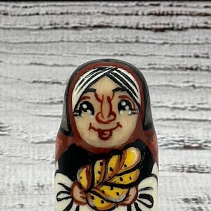 May include: A hand-painted wooden Matryoshka doll depicting a smiling woman in a headscarf, holding a loaf of bread. The doll features a black, red, and white color scheme, with a detailed facial expression and a rustic aesthetic.