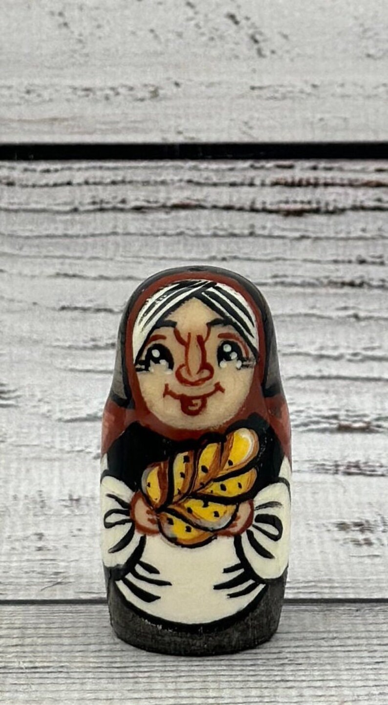 May include: A hand-painted wooden Matryoshka doll depicting a smiling woman in a headscarf, holding a loaf of bread. The doll features a black, red, and white color scheme, with a detailed facial expression and a rustic aesthetic.