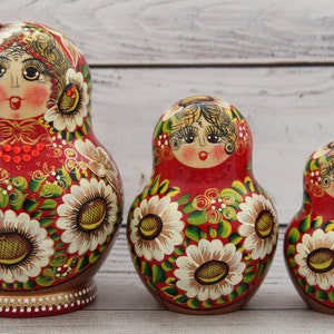 May include: Three Russian nesting dolls, or Matryoshka dolls, are displayed. The dolls are painted red with white sunflower designs and green leaves. The dolls have a woman's face on the front. The largest doll is approximately 6 inches tall.