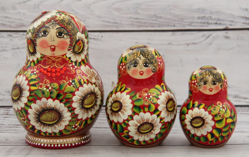May include: Three Russian nesting dolls, or Matryoshka dolls, are displayed. The dolls are painted red with white sunflower designs and green leaves. The dolls have a woman's face on the front. The largest doll is approximately 6 inches tall.