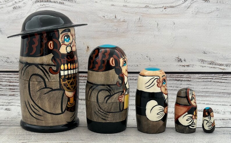 May include: A set of five wooden nesting dolls, each depicting a figure with traditional attire. The largest doll features a man with a black hat and a menorah. The dolls are painted in shades of brown, black, and beige.
