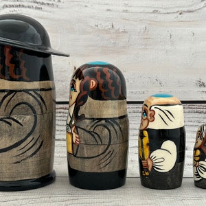 May include: A set of five wooden nesting dolls, each painted with a different character. The dolls are primarily black, brown, and beige, with details in blue, yellow, and white. The largest doll has a black hat.