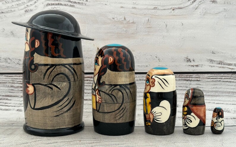 May include: A set of five wooden nesting dolls, each painted with a different character. The dolls are primarily black, brown, and beige, with details in blue, yellow, and white. The largest doll has a black hat.