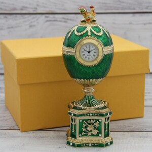 May include: An egg-shaped clock with a textured emerald green finish and gold-tone accents. The clock face is white with black Roman numerals. A small rooster figurine sits on top of the egg, which rests on a square, ornate green and gold base.