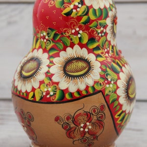 May include: A hand-painted Russian nesting doll, or Matryoshka doll, with a red background and floral design. The doll features large white sunflowers with brown centers, green leaves, and gold accents. The base is brown with red floral details.
