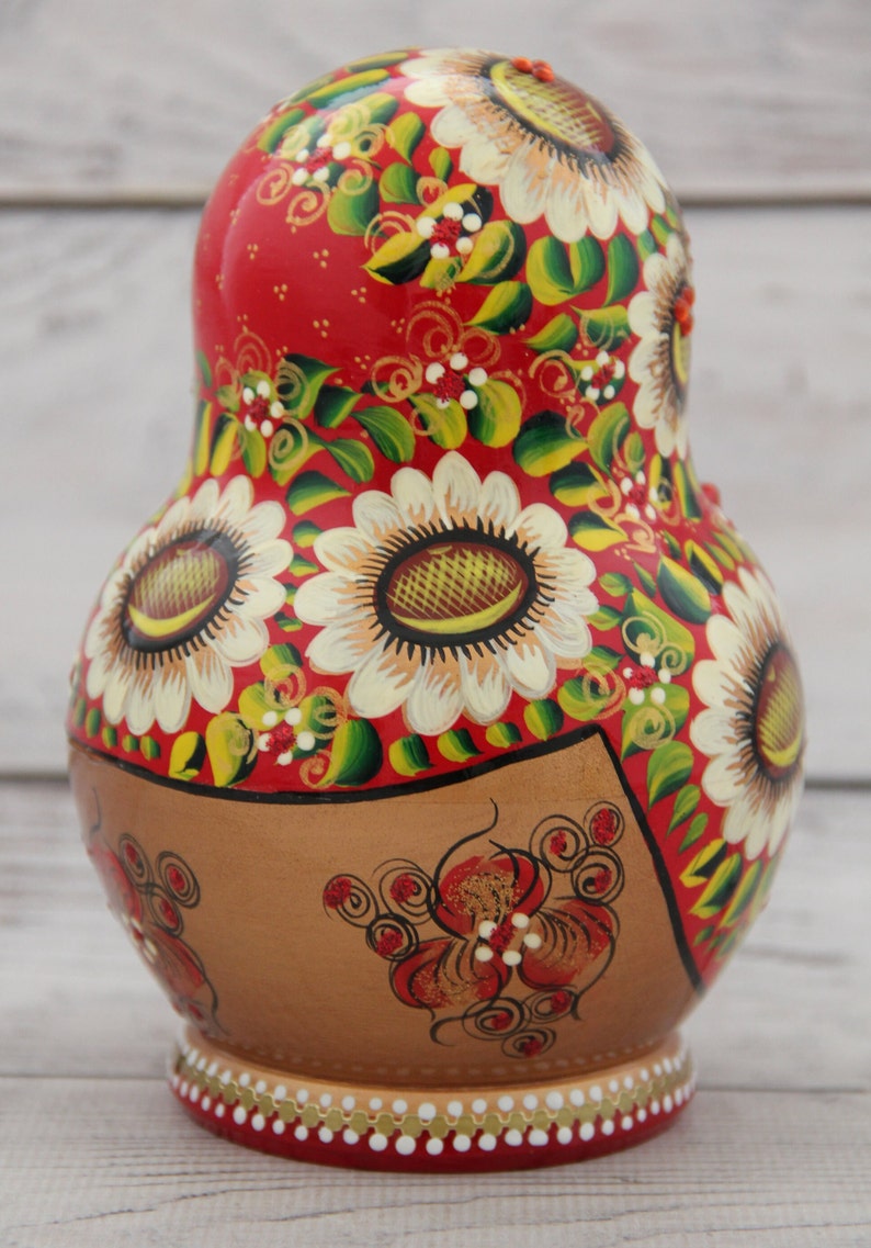 May include: A hand-painted Russian nesting doll, or Matryoshka doll, with a red background and floral design. The doll features large white sunflowers with brown centers, green leaves, and gold accents. The base is brown with red floral details.